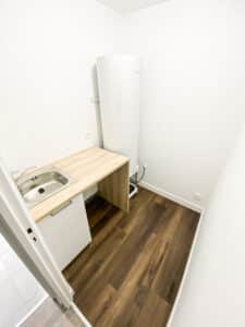Buanderie compacte avec chauffe-eau Atlantic, évier inox sur plan de travail en bois clair, meuble bas. Sol stratifié foncé, murs blancs.
