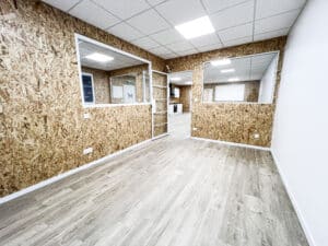Espace commercial vide, murs en panneaux OSB, plancher stratifié clair, plafond quadrillé. Fenêtres intérieures et petite kitchenette équipée.
