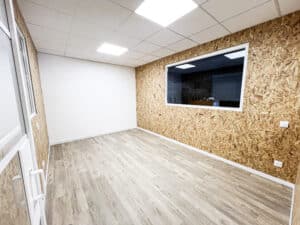Bureau moderne vide, murs OSB et blanc, sol stratifié clair. Plafond avec spots LED. Fenêtre intérieure, prises électriques.