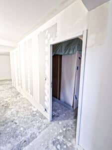 Intérieur en construction: murs en placo brut enduits, sol béton poussiéreux. Une porte ouvre sur un espace protégé par bâche verte.