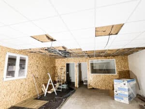 Salle en rénovation: plafond suspendu blanc démonté, isolation et câbles visibles. Murs en OSB, sol en béton. Échelles, cartons EUR ACOUSTIC.