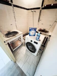 Lave-linge Samsung neuf sur carrelage moderne dans buanderie/salle de bain abîmée. Deux vieux éviers, douche. Produits XTRA et Lenor.