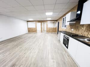 Local commercial vide avec kitchenette moderne, murs OSB, parquet clair et éclairage LED. Plafond à dalles. Signe "SORTIE" visible.