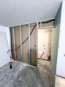 Mur en construction avec ossature métallique, câbles électriques noirs et tuyaux rouges/bleus apparents. Sol béton brut, plaques de plâtre prêtes à poser.