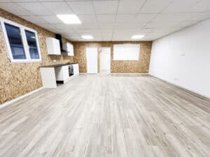 Grande pièce vide lumineuse. Murs en panneaux OSB, sol stratifié clair, kitchenette blanche, fenêtres et portes. Éclairage LED au plafond.