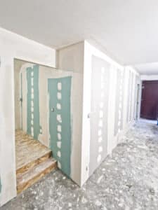 Intérieur en rénovation avec murs en placo vert et gris, partiellement enduits. Un escalier en bois OSB mène à un niveau supérieur. Sol en béton brut.