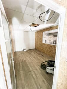 Pièce en rénovation : murs blancs et OSB, plafond suspendu, sol stratifié. Aspirateur Festool près d'une fenêtre intérieure. Travaux en cours.