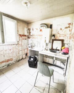 Pièce en rénovation avec murs à vif, carrelage clair, chauffe-eau, évier, miroir et outils. Chaise grise, travaux en cours.
