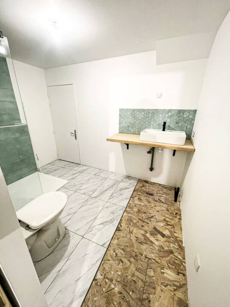 Salle de bain en rénovation avec moitié du sol carrelée blanc marbré, toilettes, et zone en OSB. Lavabo moderne sur planche bois, crédence hexagone vert.