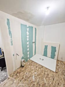 Salle de bain en rénovation avec receveur de douche blanc, murs en placo vert, sol OSB et plafond taché par l'eau.