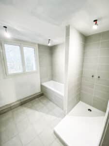 Salle de bain moderne en rénovation avec carrelage mural et au sol gris, baignoire blanche et receveur de douche intégré.