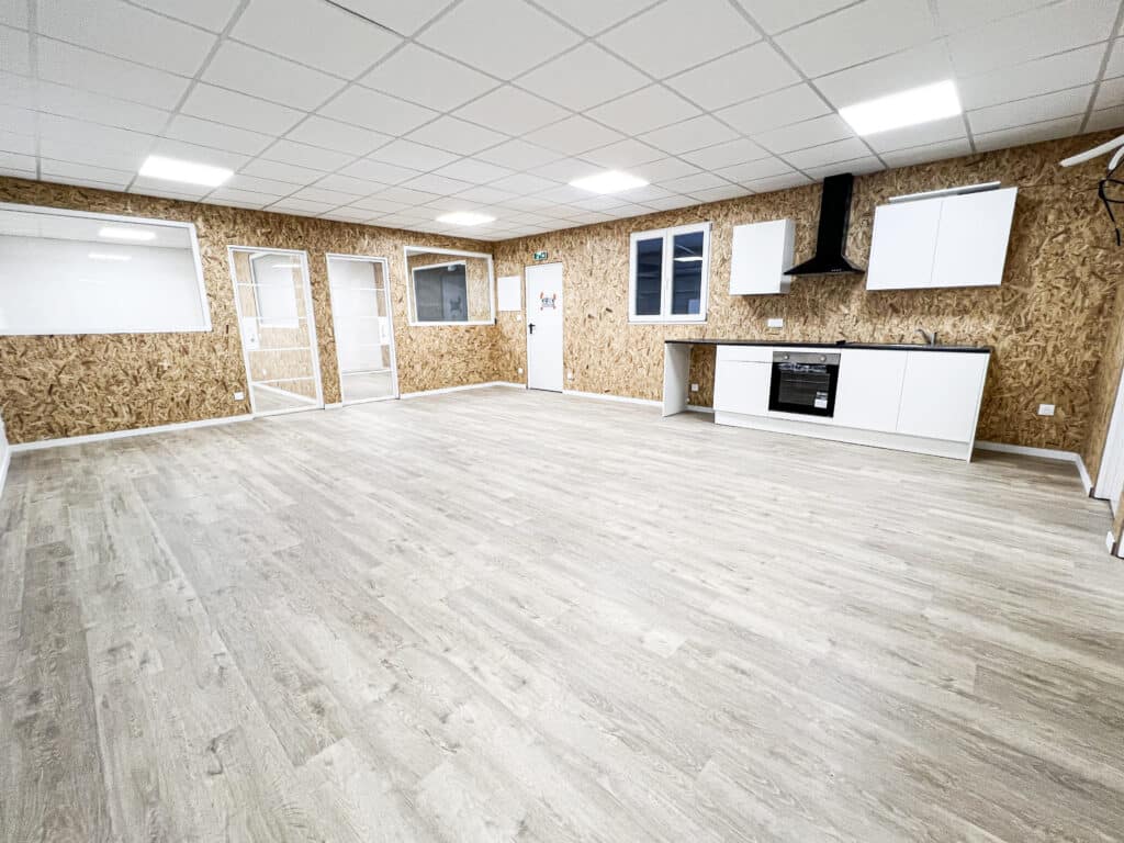 Salle lumineuse avec murs en panneaux OSB, sol stratifié clair et cuisine moderne blanche équipée d'un four. Portes vitrées et enseigne EXIT.
