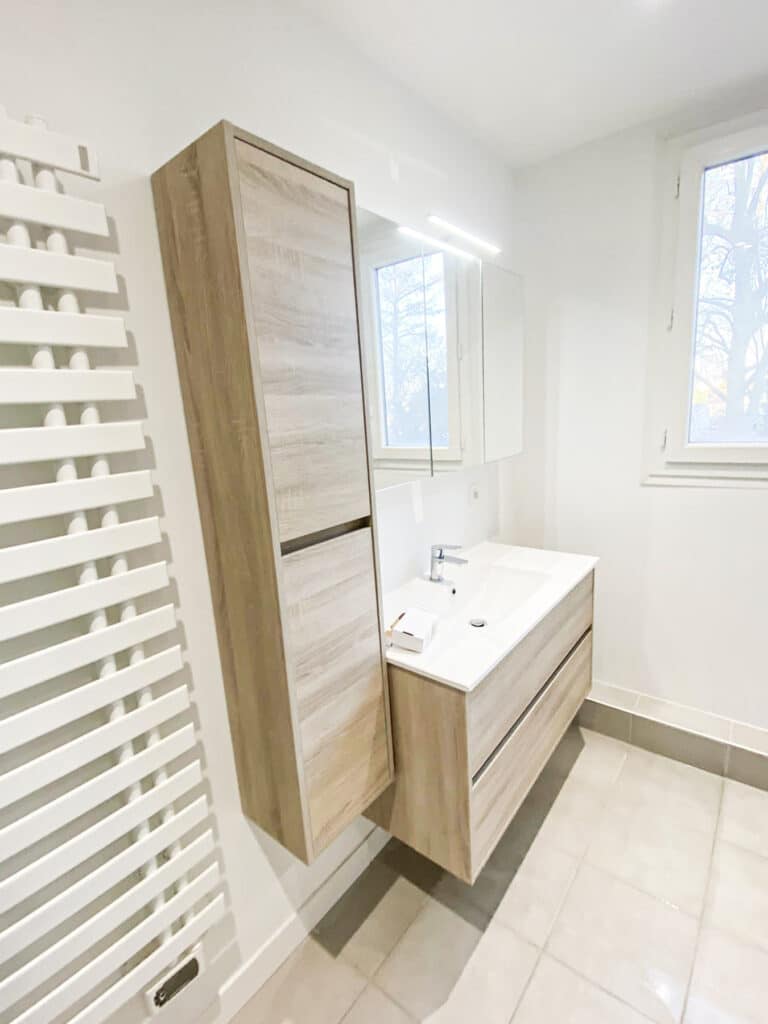 Salle de bain moderne: meuble vasque et colonne de rangement murale en bois clair, miroir lumineux, sèche-serviettes blanc.