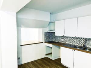 Cuisine moderne: meubles blancs, plans de travail bois, crédence à motifs bleus/blancs. Évier double, plaque cuisson, hotte, passe-plat. Sol parquet.