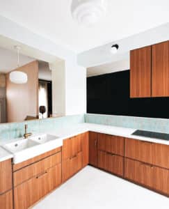 Cuisine design aux meubles bois clair, plan travail blanc, double évier laiton. Crédiance turquoise, vue sur salon pêche et suspension.