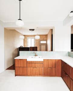 Cuisine lumineuse avec îlot central en bois clair et comptoir blanc, double évier de ferme, robinet laiton. Crédence carrelage turquoise, murs pierre et plafonds blancs.