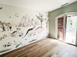 Chambre d'enfant lumineuse avec fresque murale de dinosaures, montagnes et arbres, parquet bois et mur vert sauge. Porte ouverte.