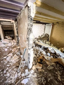 Intérieur en pleine démolition, murs partiellement ouverts, gravats partout. Poutres apparentes violettes et jaunes. Ambiance de chantier de rénovation.