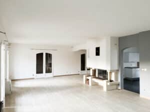 Grand salon vide, lumineux et rénové avec parquet clair. Cheminée moderne, portes-fenêtres, accès cuisine. Prêt à être aménagé.