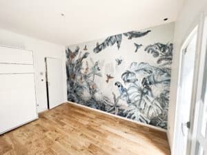 Murale tropicale vive avec perroquet bleu, oiseaux, papillons et feuillage luxuriant sur un mur blanc. Sol en parquet clair, placard blanc moderne et porte-fenêtre.