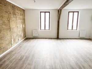 Pièce vide lumineuse avec mur en pierre apparent, murs blancs, parquet clair, deux fenêtres et radiateurs, poutre bois apparente.