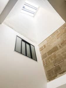 Intérieur lumineux avec puits de lumière. Murs contrastés : blanc lisse, texturé, et briques de pierre apparentes. Fenêtre moderne.