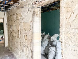 Couloir en rénovation avec murs en pierre brute et plafond exposé, câbles électriques bleus. Une pièce adjacente contient des sacs de débris blancs.