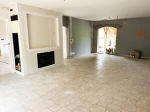 Pièce en rénovation avec cheminée moderne encastrée, niche à bois, et murs gris inachevés. Sol en carreaux hexagonaux beiges. Sacs TOUPRET.