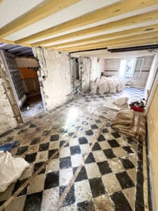 Pièce en démolition avec carrelage à motifs d'échiquier noir et beige, murs abattus, poutres apparentes au plafond et sacs de gravats.