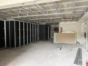 Intérieur en rénovation avec ossature métallique au plafond et murs isolés en noir. Plaques de placo, profilés métalliques et extincteur rouge au sol.