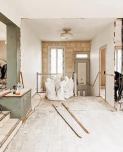 Intérieur d'une maison en rénovation, murs en partie démolis. Sol brut, sacs de gravats blancs et outils. Deux ouvriers travaillent près d'un escalier et de fenêtres.
