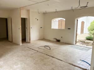 Pièce en cours de rénovation, murs blancs bruts, fils électriques au plafond, carrelage beige au sol, tranchée, tuyaux de plomberie, fenêtre et portes ouvertes.