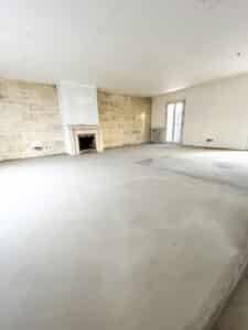 Pièce vide en rénovation avec un mur en pierres apparentes et une cheminée. Le sol en béton clair est brut et une fenêtre est visible.