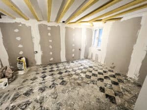 Pièce en rénovation : murs placo enduits, poutres apparentes au plafond. Vieux carrelage à échiquier noir et blanc. Sac "MIP" au sol. Fenêtre.