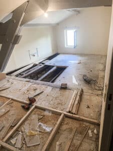 Pièce en rénovation avec plancher partiellement démoli, poutres en bois apparentes, outils de chantier et gravats. Plafond mansardé et fenêtre. Travaux en cours.