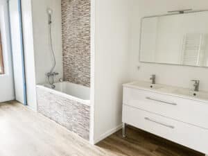 Salle de bain moderne blanche avec baignoire carrelage mosaïque marron, meuble double vasque sous grand miroir éclairé. Sol aspect bois clair.
