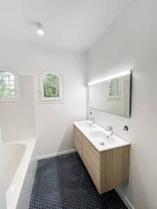Salle de bain moderne: double vasque bois, miroir lumineux, carrelage hexagonal bleu, baignoire et fenêtres cintrées avec verdure.