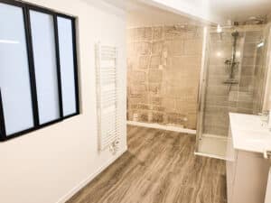 Salle de bain moderne: mur en pierre apparente, douche vitrée, lavabo blanc, radiateur sèche-serviettes, fenêtre noire, parquet bois.