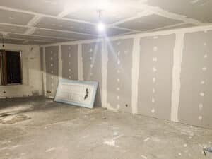 Intérieur en travaux: murs et plafond en plaques de plâtre avec joints enduits. Une ampoule éclaire le sol brut et une porte neuve est posée.