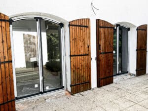 Façade blanche avec trois portes coulissantes en verre à arcades, flanquées de volets en bois rustiques partiellement ouverts sur une terrasse carrelée.